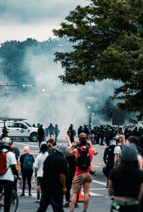 Personen beteiligen sich an einem Protest, Tränengas wird im Hintergrund eingesetzt. 