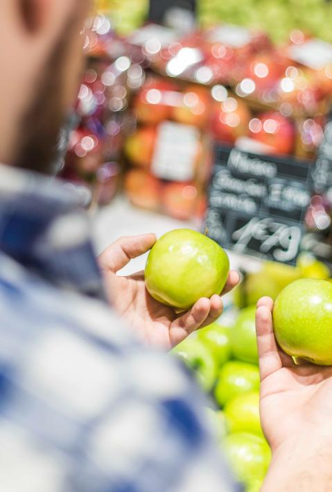 Ein Mann beim Einkauf, er betrachtet zwei grüne Äpfel in seinen Händen. 