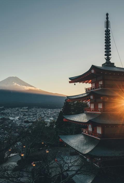 A picturesque scene featuring a traditional Japanese pagoda with multiple tiers, bathed in the warm light of the setting sun. In the background, the majestic Mount Fuji rises, partially covered in snow and surrounded by a layer of clouds. The foreground includes a town nestled at the base of the mountain, with trees and lanterns lining a path leading towards the pagoda. The sky is clear, adding to the serene and tranquil atmosphere of the image.