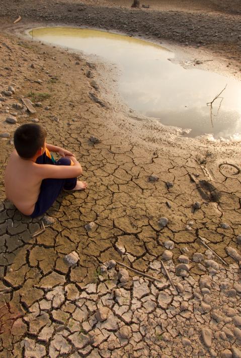 Ein Kind sitzt auf ausgetrockneter, zerrissener Erde und schaut auf eine schrumpfende Wasserlache.