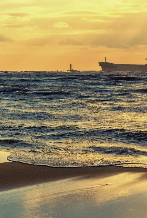 Cargo ships are in the Baltic sea at sunset past the lighthouse against the sky with sun rays