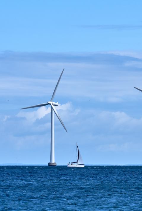 Three wind mills standing offshore at sea near Amager Beach Park in Denmark, with a sail boat sailing in front