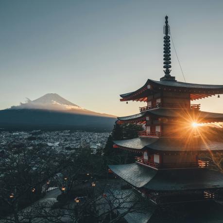 A picturesque scene featuring a traditional Japanese pagoda with multiple tiers, bathed in the warm light of the setting sun. In the background, the majestic Mount Fuji rises, partially covered in snow and surrounded by a layer of clouds. The foreground includes a town nestled at the base of the mountain, with trees and lanterns lining a path leading towards the pagoda. The sky is clear, adding to the serene and tranquil atmosphere of the image.
