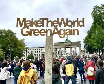 Demo am Brandenburger Tor mit einem Stock im Vordergrund, das Buchstaben balanciert, die folgenden Satz bilden: "Make The World Green Again". 