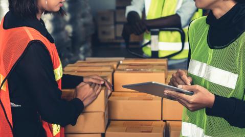 Two workers in yellow jackets are standing next to packaged goods and discuss logistics