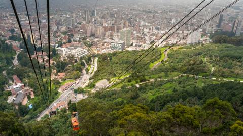 aerial view from Monserrate on top of the mountain where you can see downtown Bogota with tall buildings and road between the woods. Below we also see the cable car of the city arriving at its base.